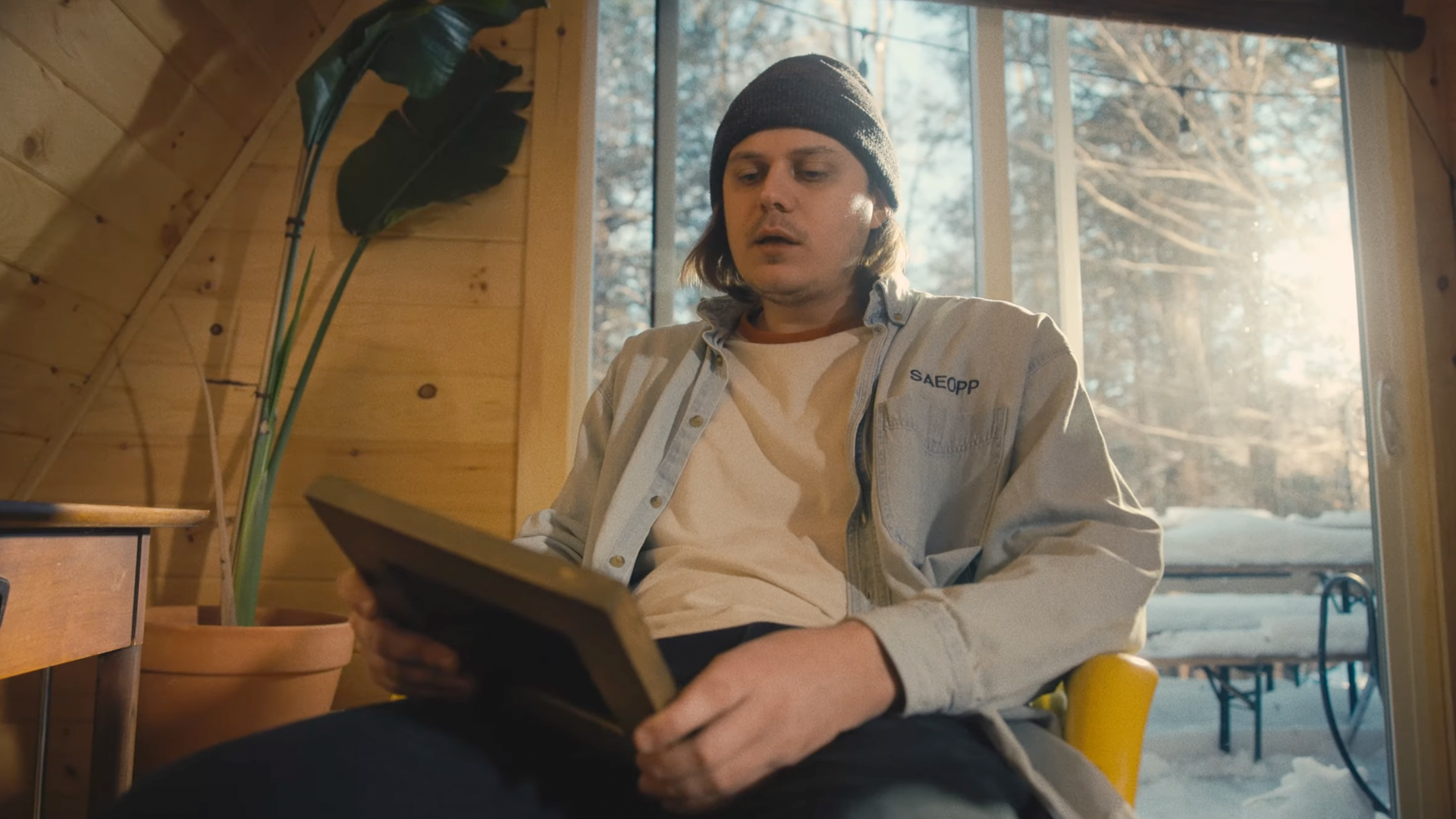 A young man with shoulder-length hair, wearing a beanie and a light-colored work shirt, sitting inside a wooden cabin reading a book or magazine, with snowy trees visible through a large window behind him.