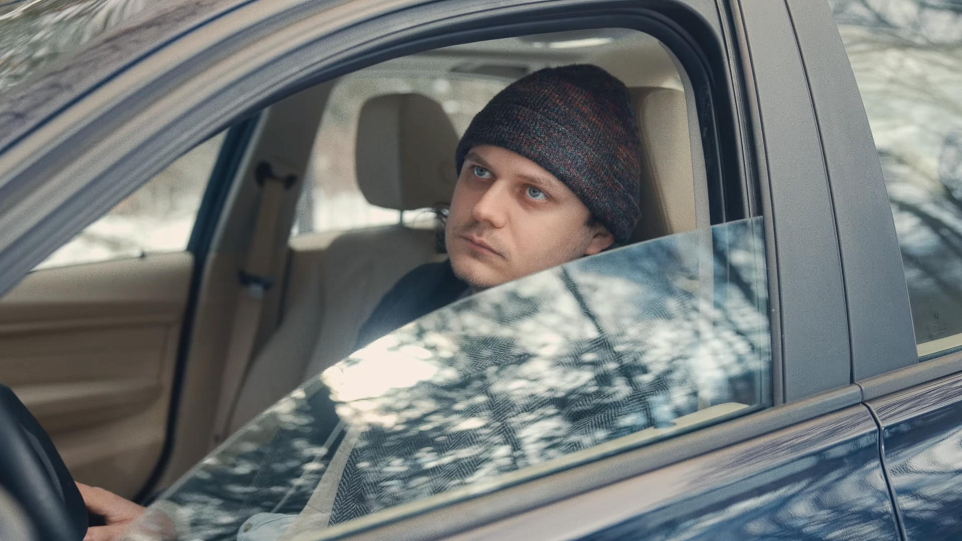A man with a beanie sitting in the front seat of a car, looking out the window with a serious expression. The car's interior is beige, and there are reflections of trees on the window.