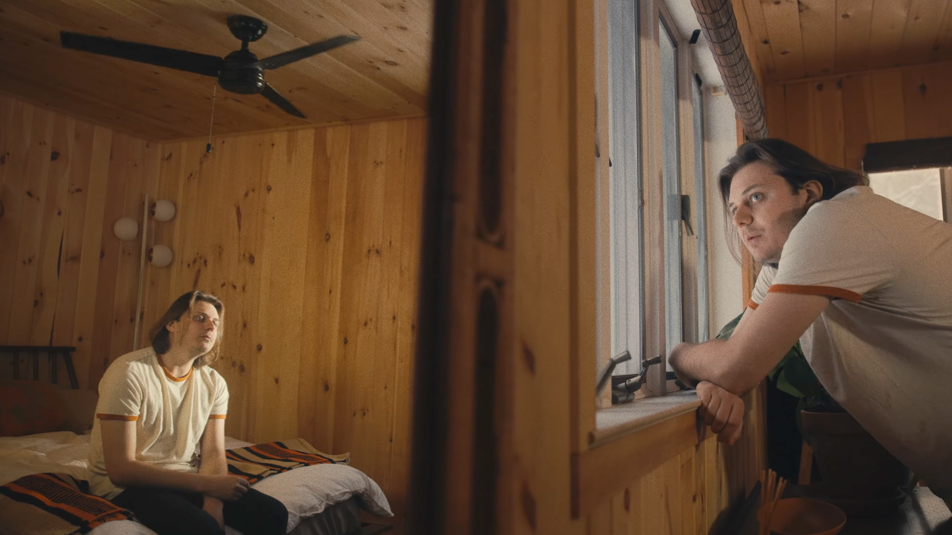 A man with shoulder-length hair and glasses sitting on a bed inside a wooden room, looking pensively out the window where a woman leans on the windowsill.