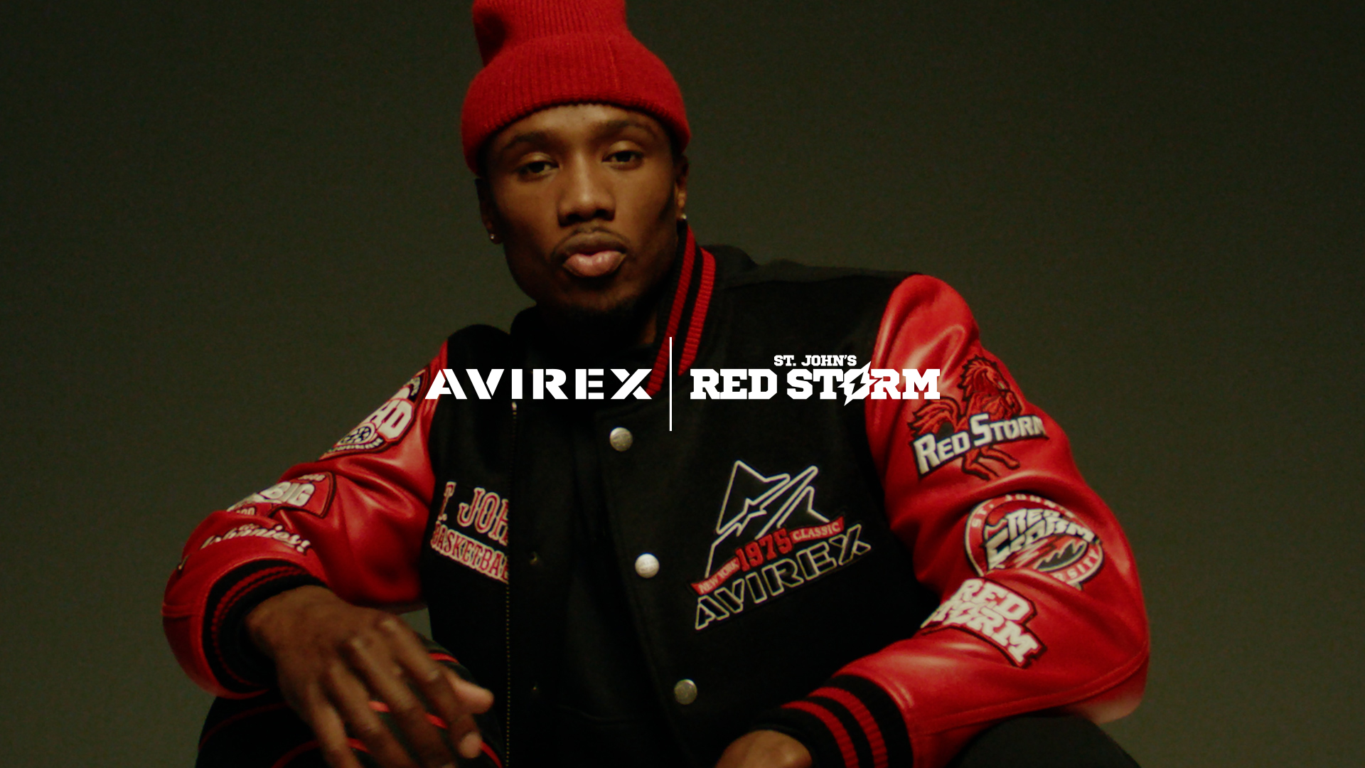 A young man wearing a red beanie and a red and black varsity jacket with patches, sitting against a plain dark background.