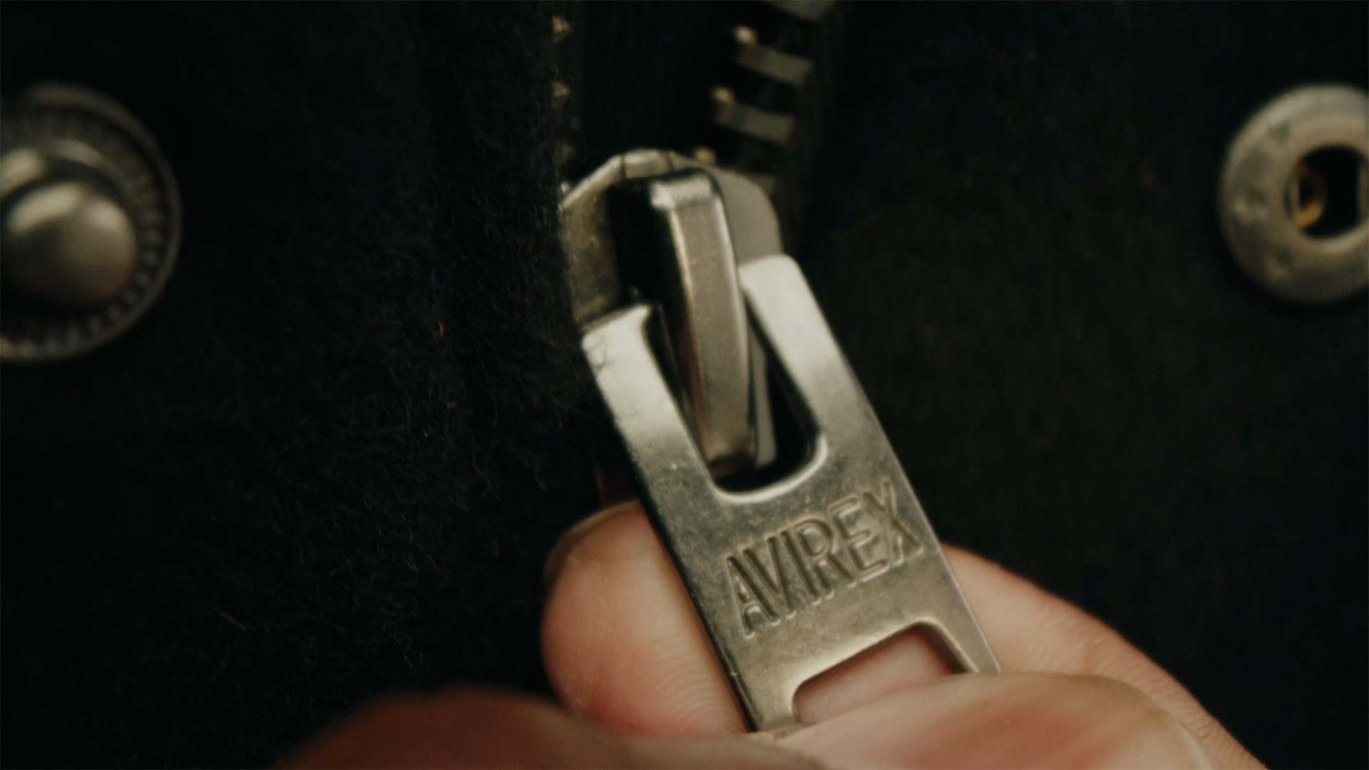 Close-up of a hand holding an Avirex branded zipper on a black fabric surface.