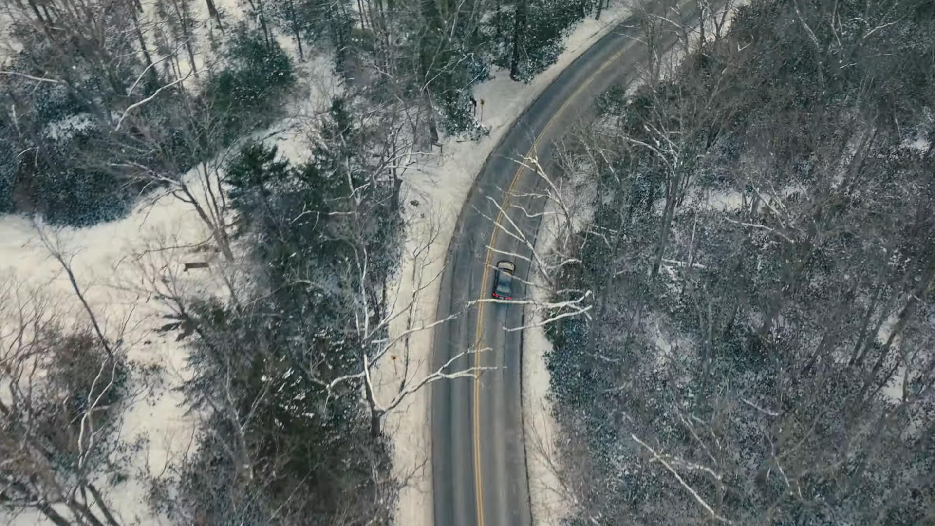 A winding two-lane road through snow-covered forest, viewed from above, with a black vehicle driving along it.