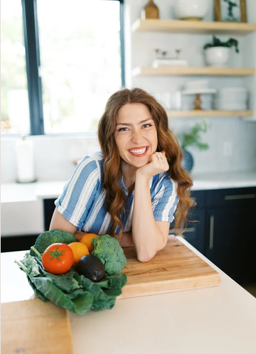 A woman with long wavy hair smiling at the camera, resting her chin on her hand in a modern kitchen with fresh vegetables on the counter.