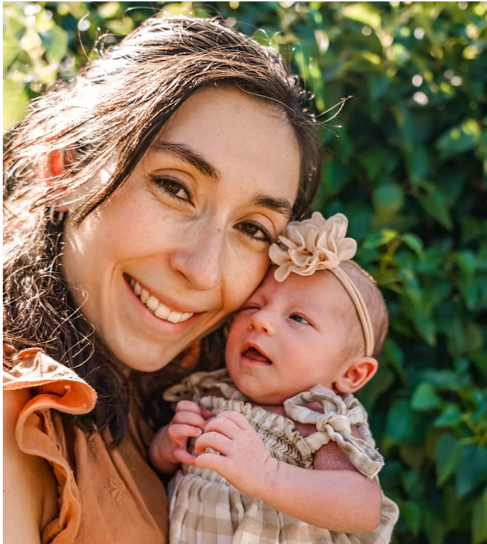A woman smiling, holding a newborn baby girl outdoors with green foliage in the background.