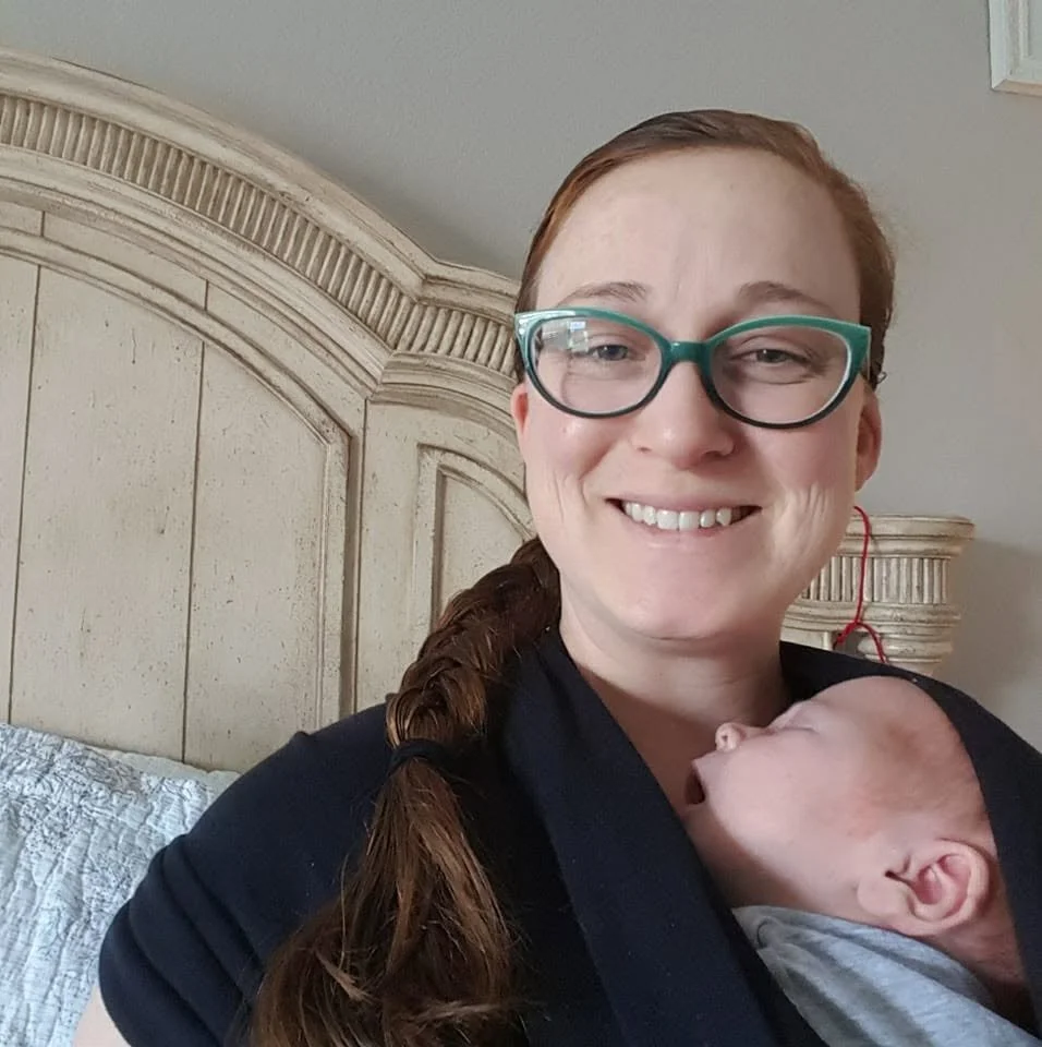 A woman with red hair, wearing glasses, smiling while holding an infant with a hand near her chest. The woman is sitting against a wooden headboard in a bedroom.
