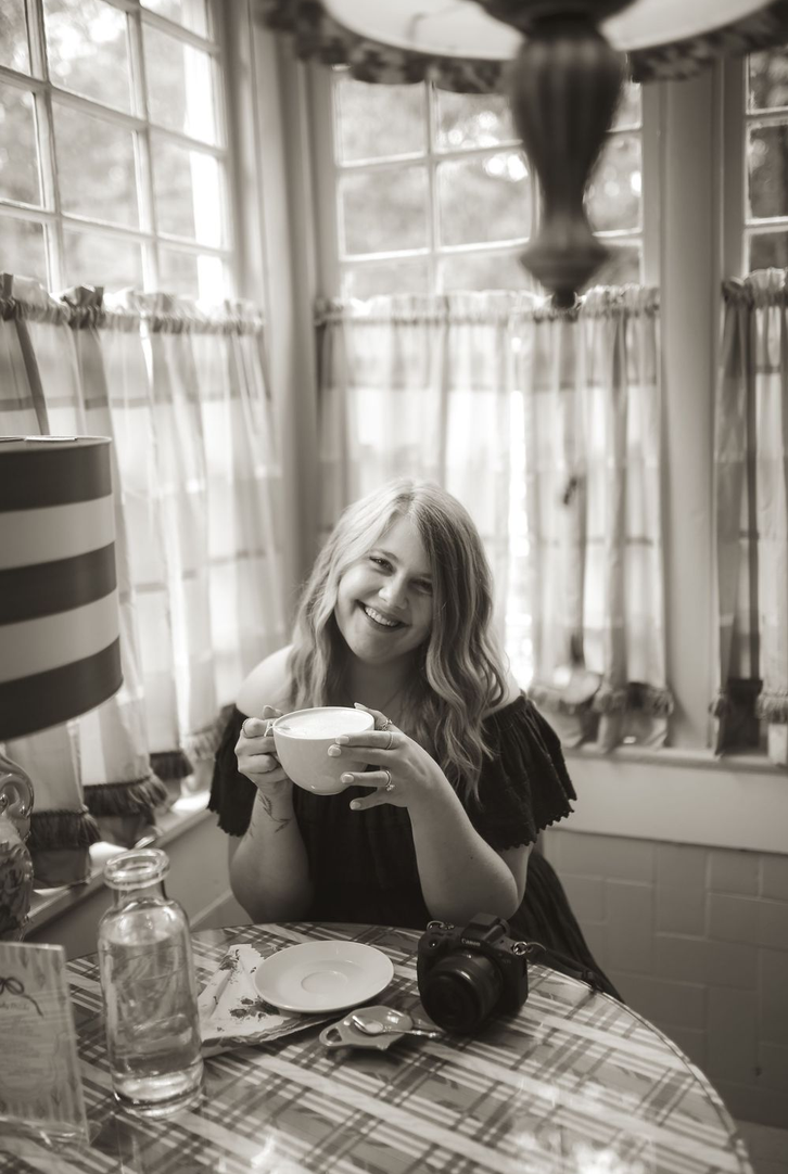 A young woman with wavy hair smiling and holding a mug, sitting at a table in a bright room with plaid tablecloth and checkered curtains.