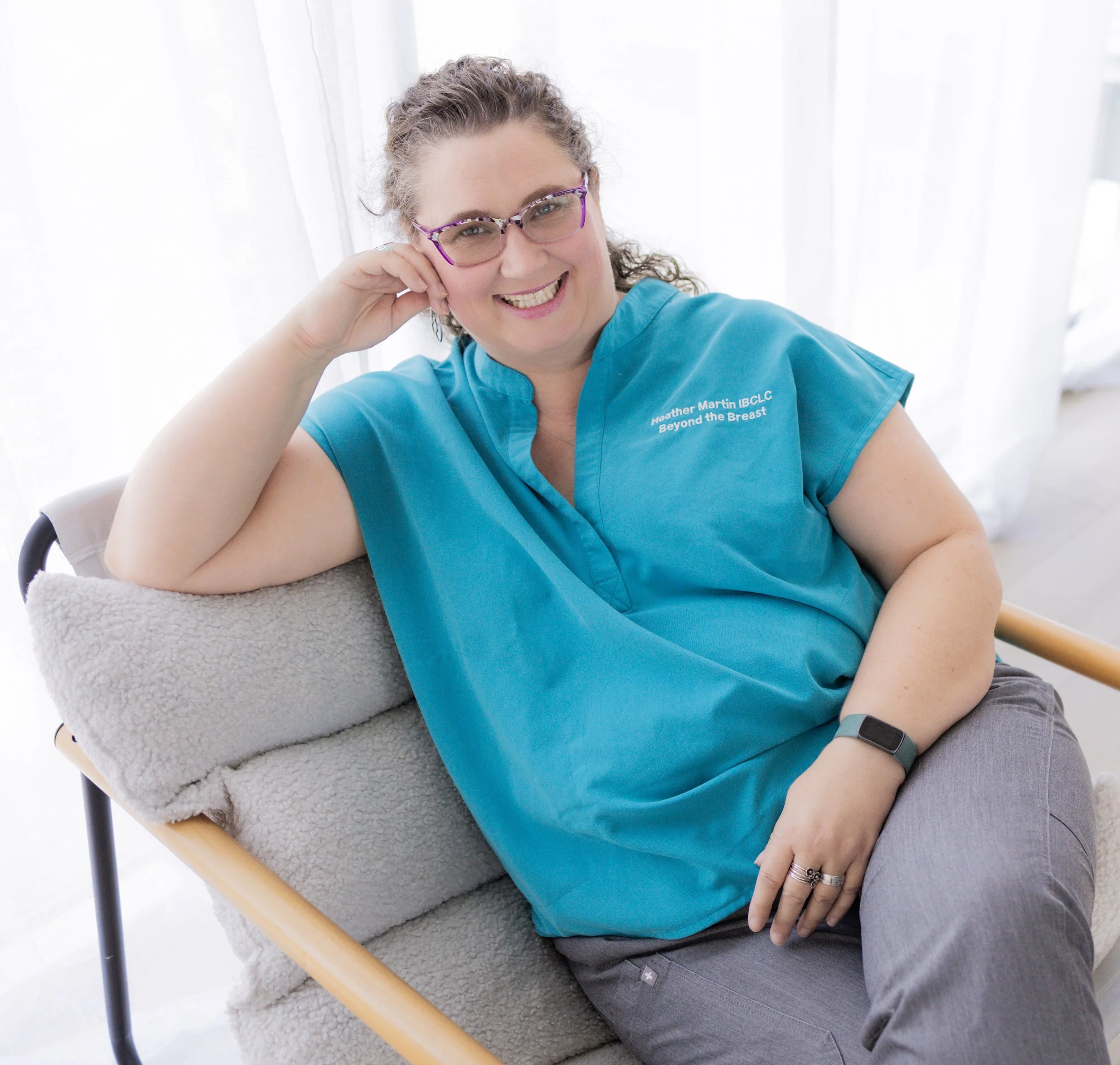 A woman with curly hair, wearing purple glasses and a teal medical uniform, smiling and sitting in a chair covered with a soft, textured blanket in front of a white background.