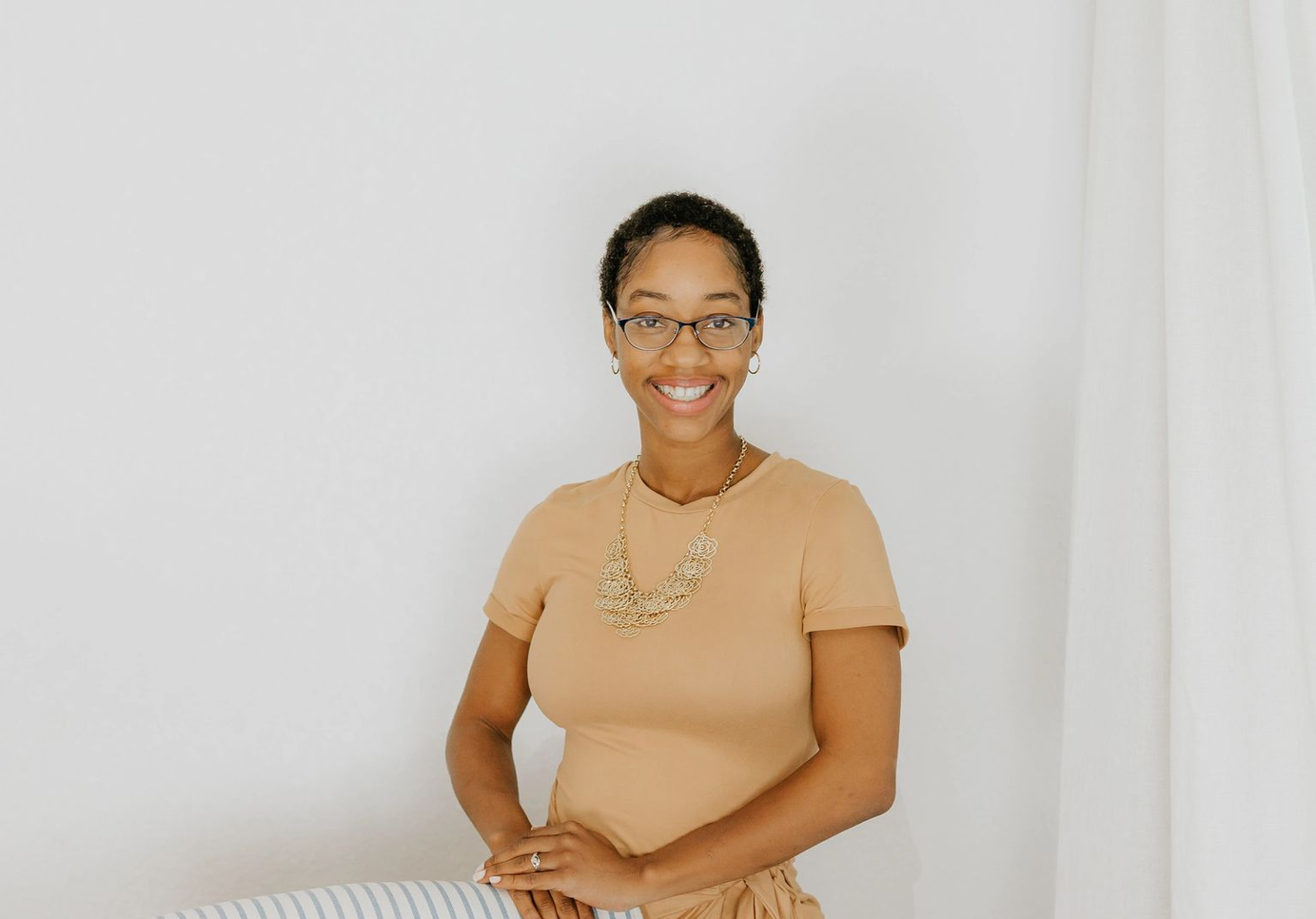 Smiling woman with glasses and short curly hair, wearing a tan shirt and gold jewelry, standing in front of a white wall with a white curtain on the right.
