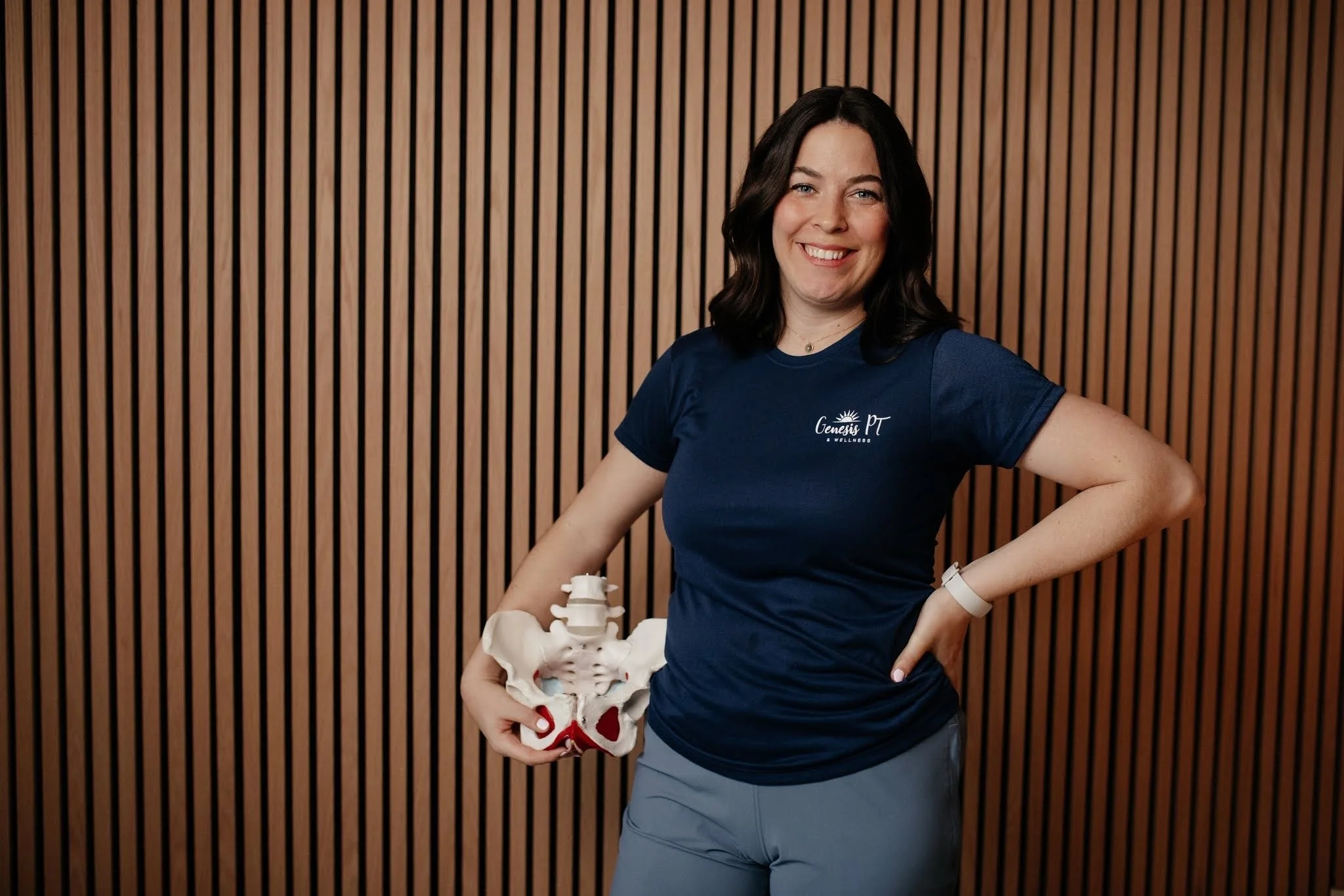 A woman wearing a Genesis PT Wellness t-shirt standing in front of a wooden slat wall, holding a model of a pelvis.