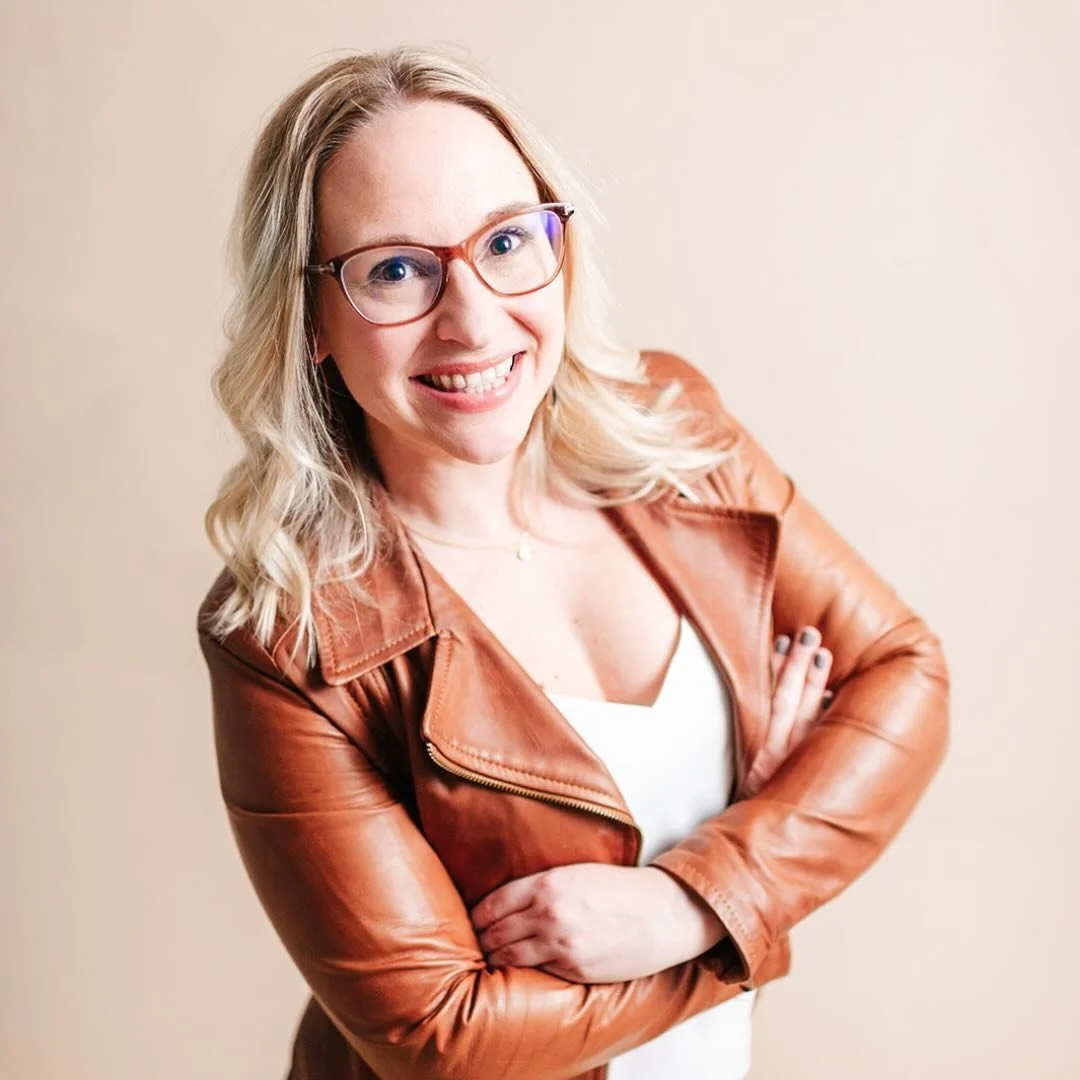 A woman with blonde hair, glasses, and a leather jacket smiling with crossed arms against a plain background.