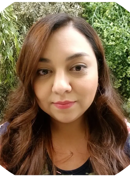 A woman with long, wavy brown hair and light makeup standing outdoors in front of green foliage.