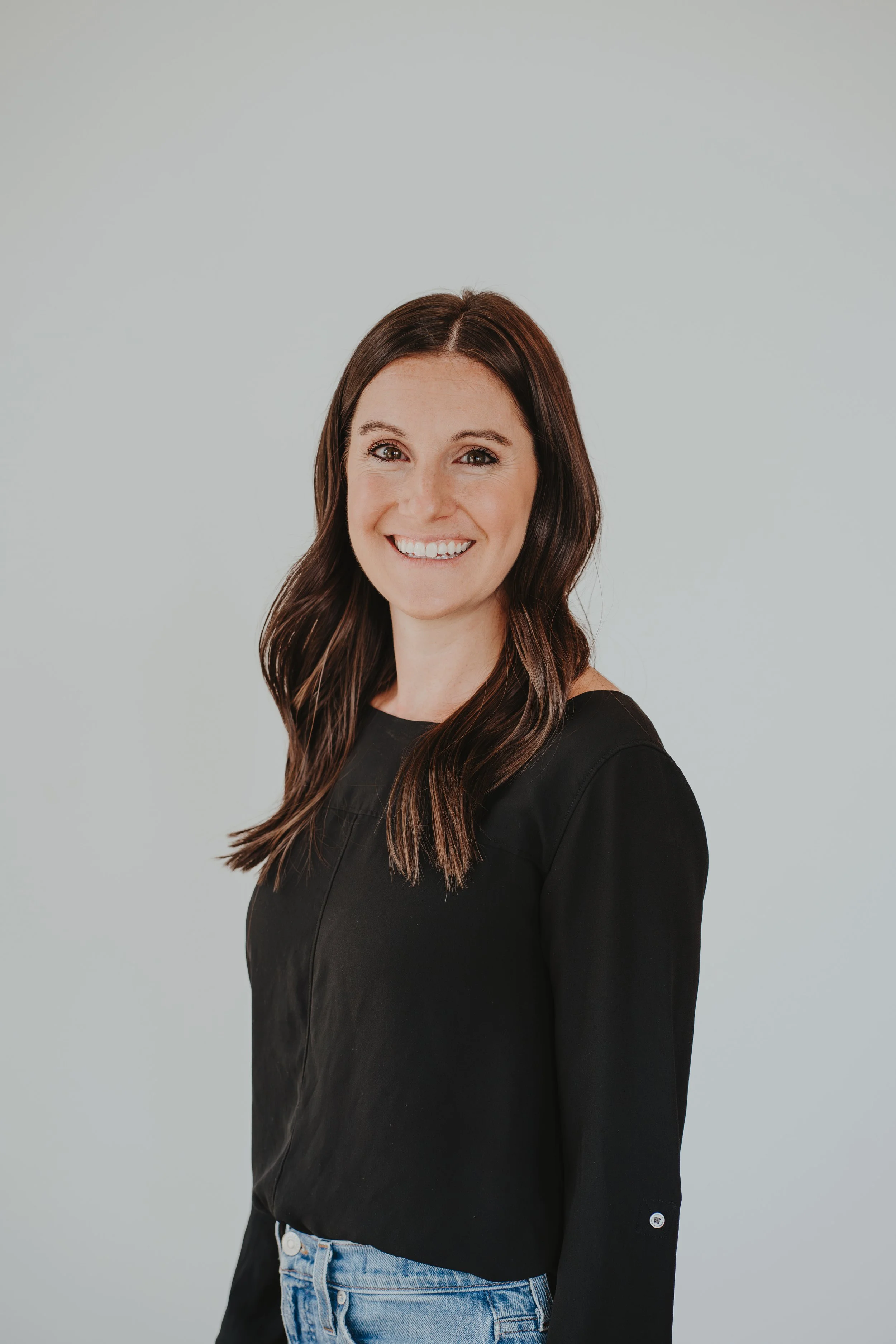 A smiling woman with shoulder-length brown hair wearing a black top and light blue jeans against a plain gray background.