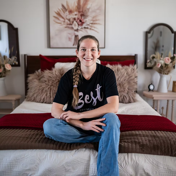 Young woman with long braided hair, wearing a black T-shirt and jeans, sitting on a bed with floral decor in the background in a cozy bedroom.