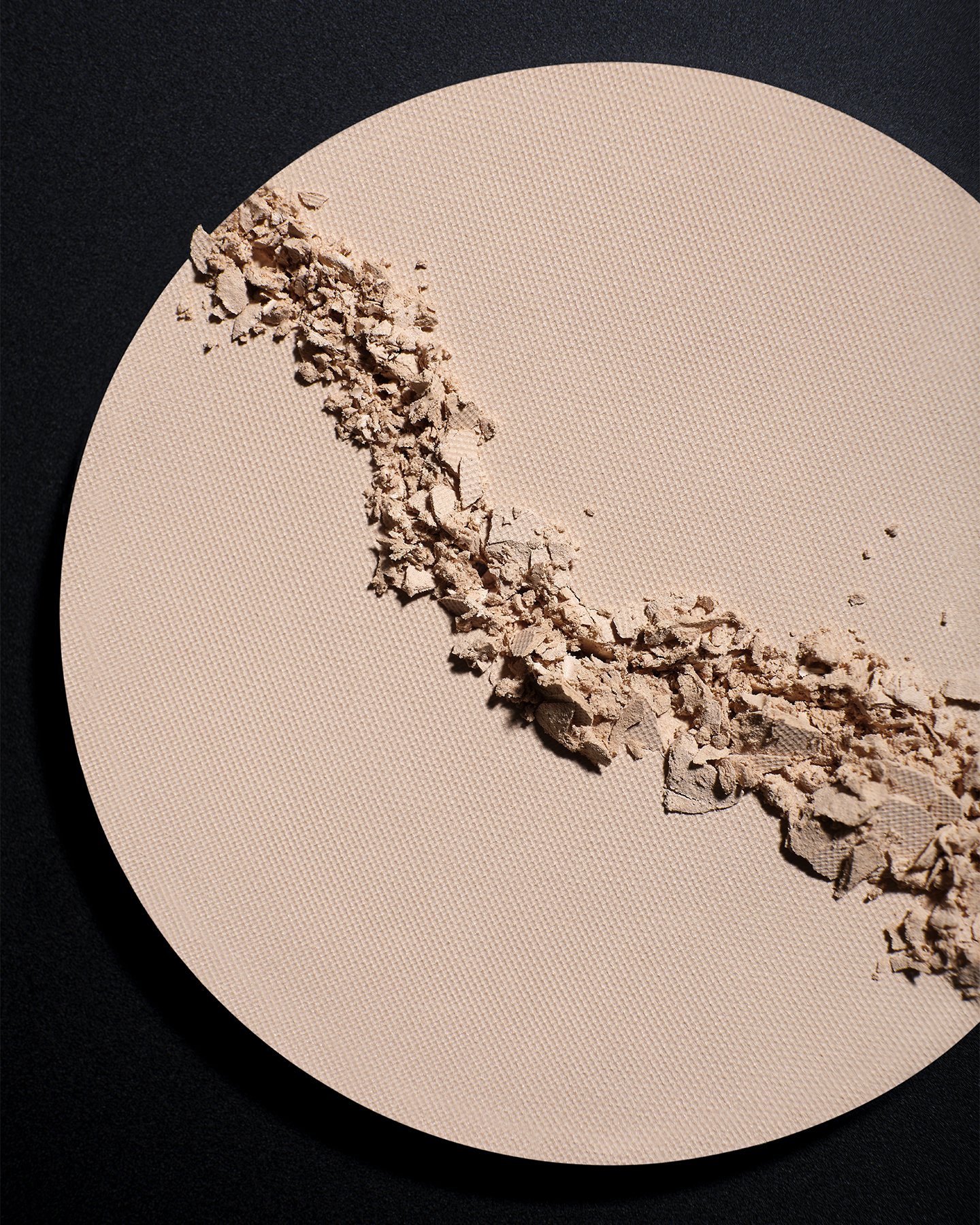 A circular beige makeup compact with crushed powder inside, on a black background.
