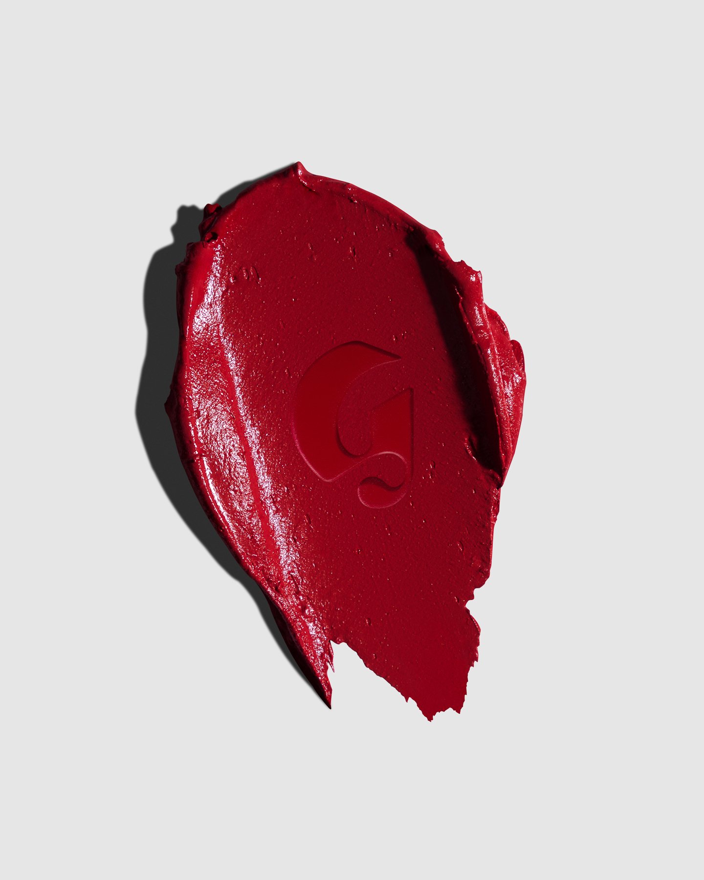 A broken red lipstick with a visible Glossier logo on a light gray background, casting a shadow.