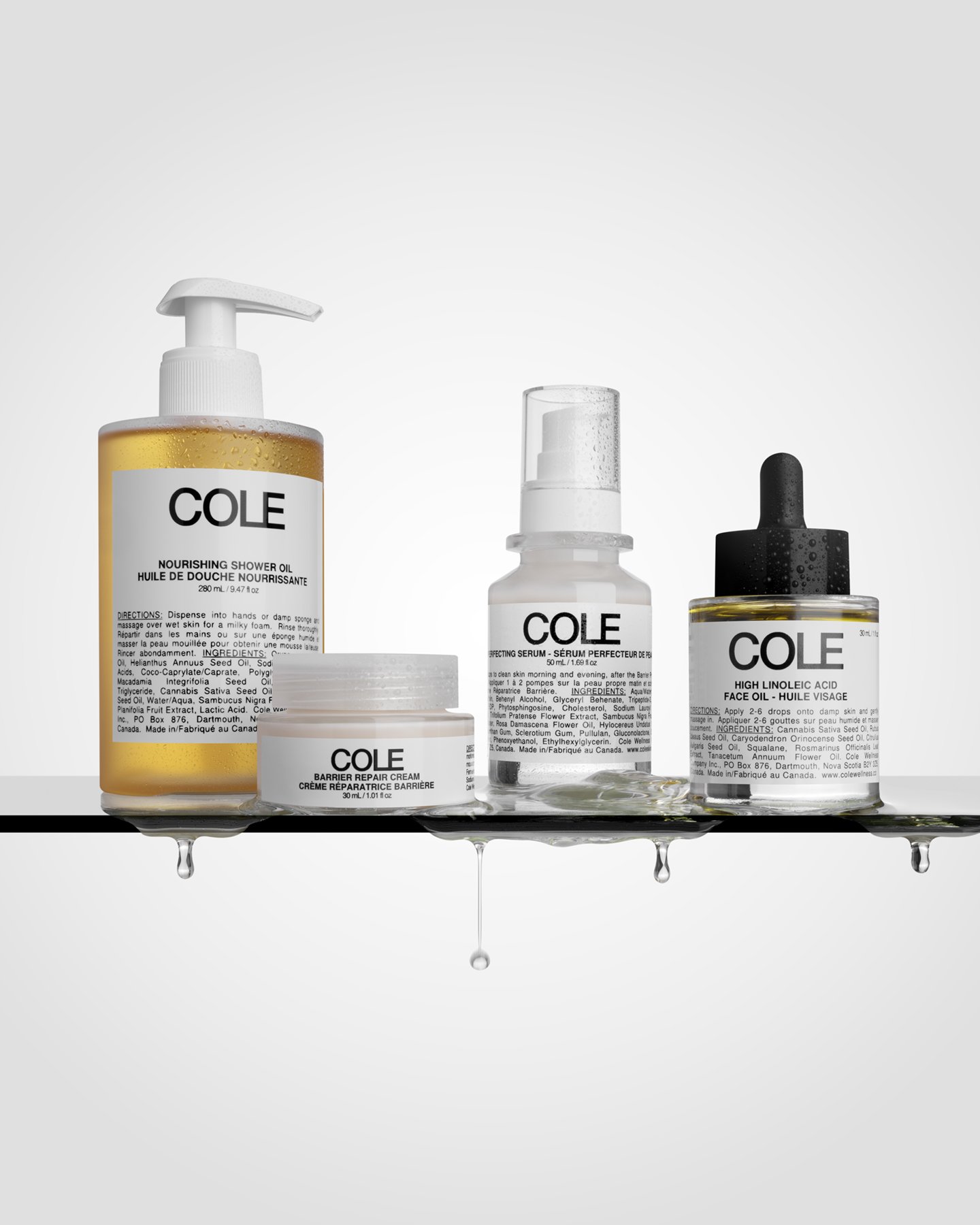 Set of skincare products on a glass shelf with water droplets, including a nourishing shower oil, barrier repair cream, facial serum, and high linoleic acid face oil, all with white labels and black text.