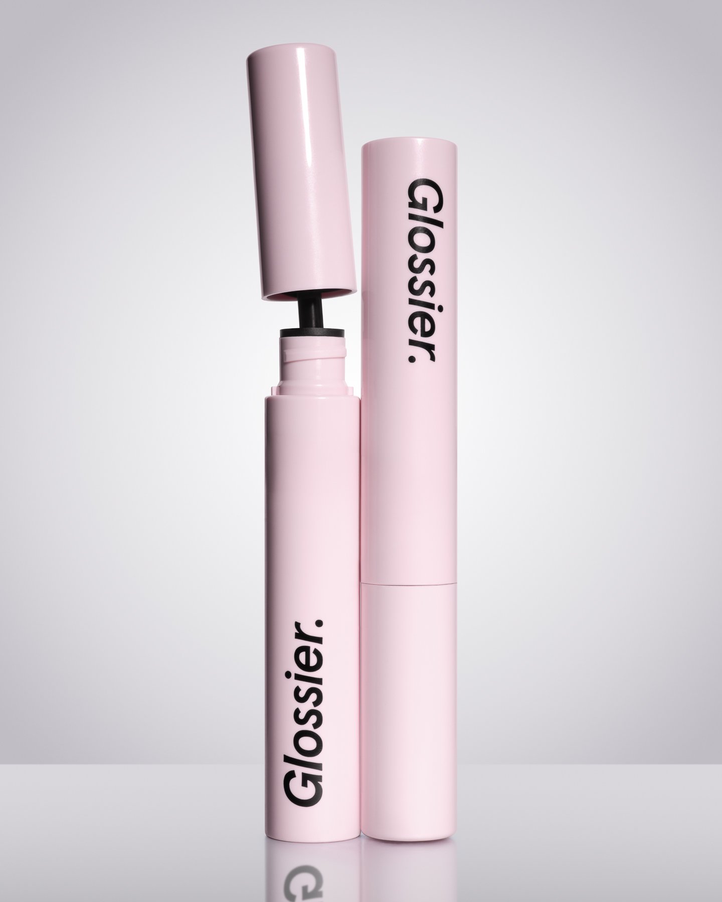 Pink lip gloss tube with the cap removed, revealing the applicator wand, standing next to a matching pink container with the brand name 'Glossier' in black letters.