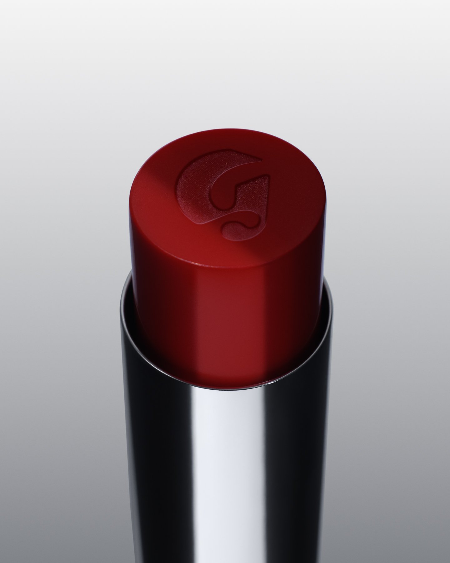 Red lipstick tube with Glossier logo engraved on the top, standing upright against a light gradient background.