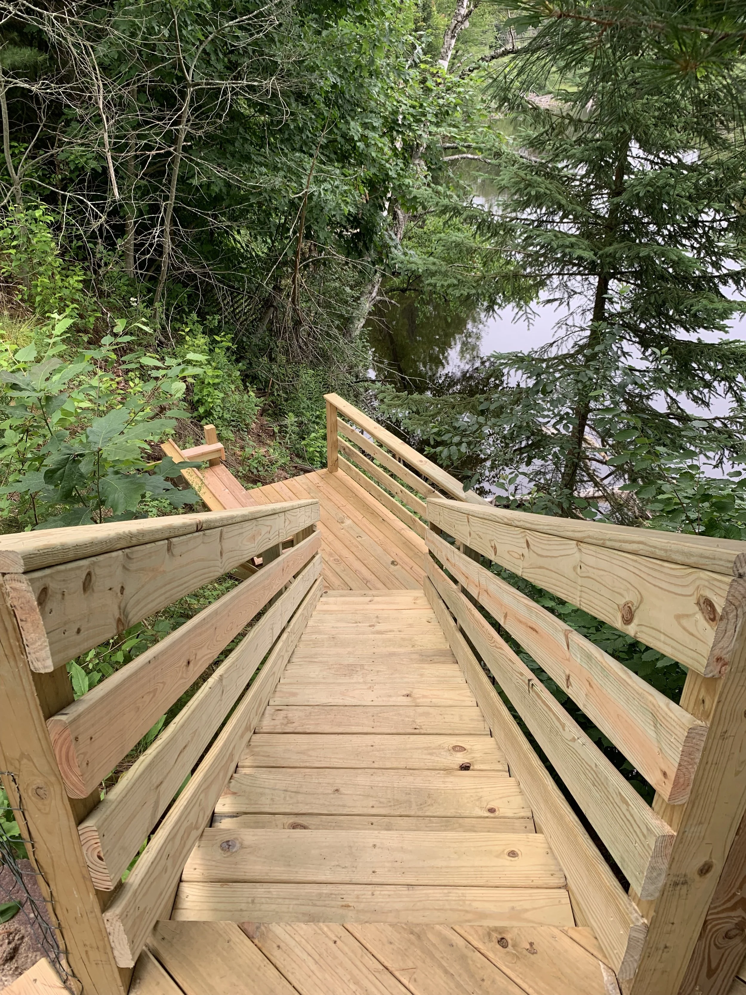 Newly built wooden staircase leading down to a forested area next to a river, surrounded by green trees and foliage.