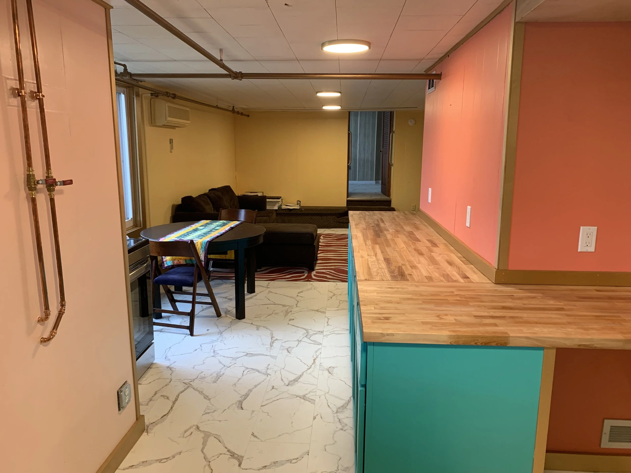 Living room with yellow walls, a brown couch, a small black dining table with a colorful table runner, and a staircase leading upstairs. The room has a mix of tiled and wooden flooring, with pink and teal accents on the walls.