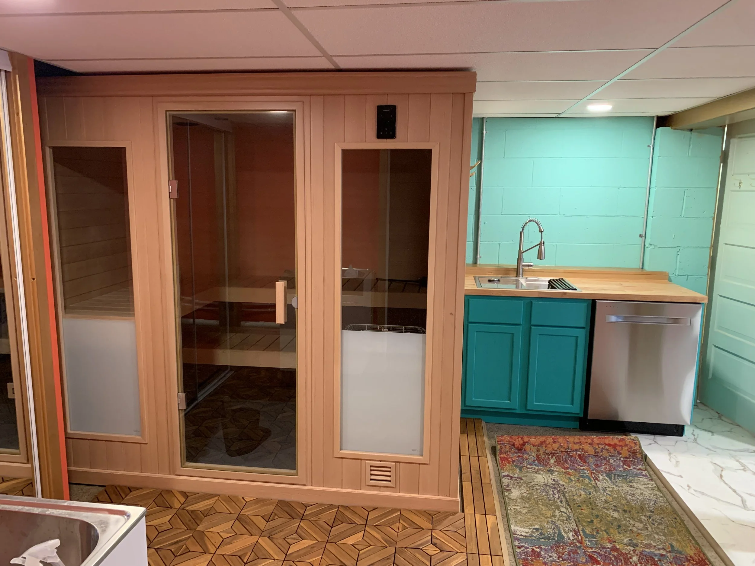 A room with a wooden sauna on the left and a small kitchenette on the right, featuring teal cabinets, a wooden countertop, a sink with a faucet, a dishwasher, and a colorful rug on a tiled floor.