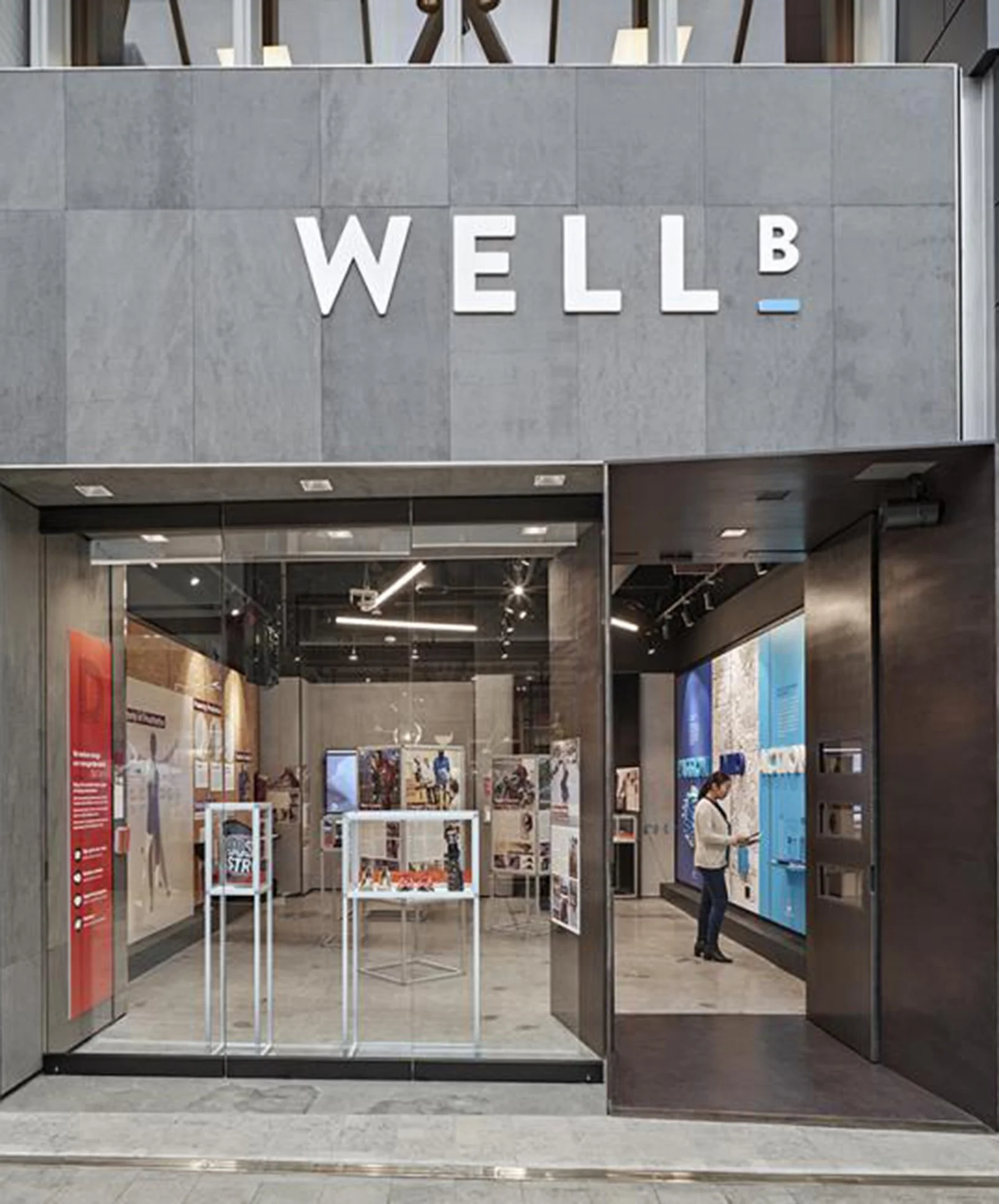 Entrance to a modern gallery or museum with large glass doors, displaying various exhibits inside and a person interacting with an information kiosk.