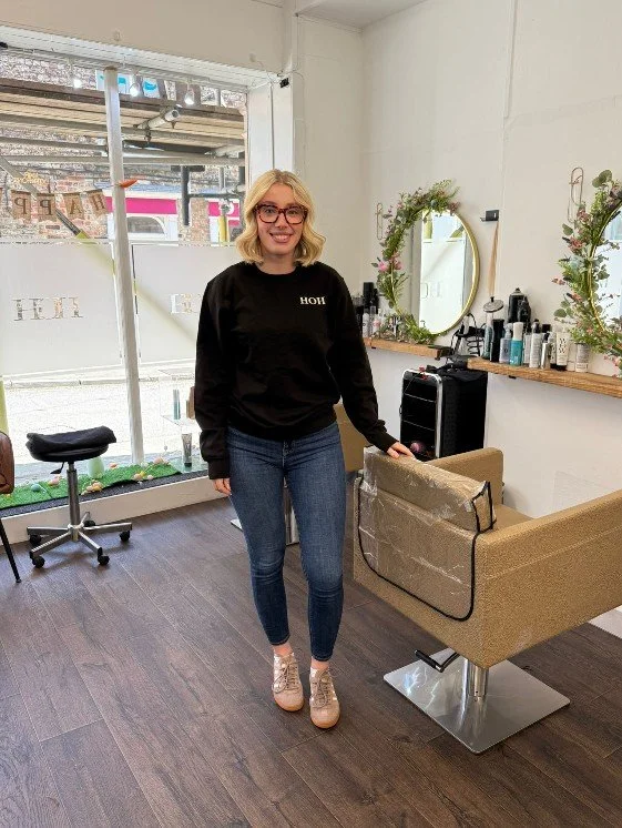 A woman with blonde hair and glasses smiling inside a hair salon, standing next to a styling chair with a protective cape, and a mirror on the wall behind her, decorated with floral arrangements.