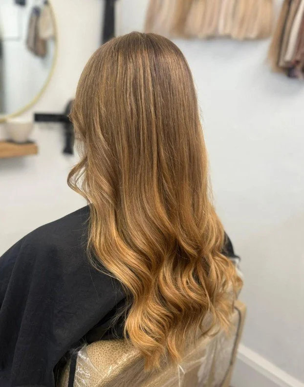 Back view of a woman with long, wavy, blonde hair sitting in a salon chair.