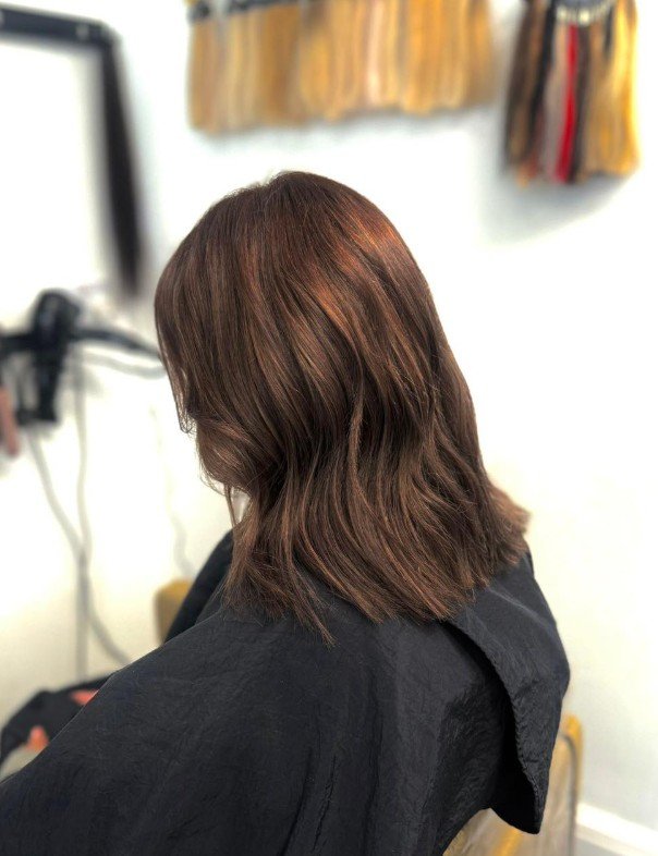Back of a woman with shoulder-length brown hair sitting in a salon chair, with shelves of hair products in the background.
