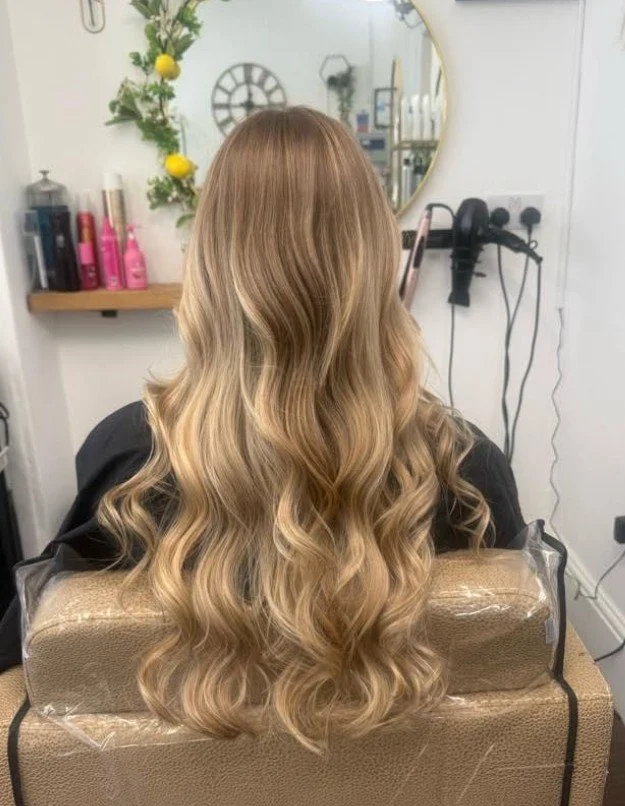 Back view of a woman with long, wavy blonde hair sitting in a salon chair.