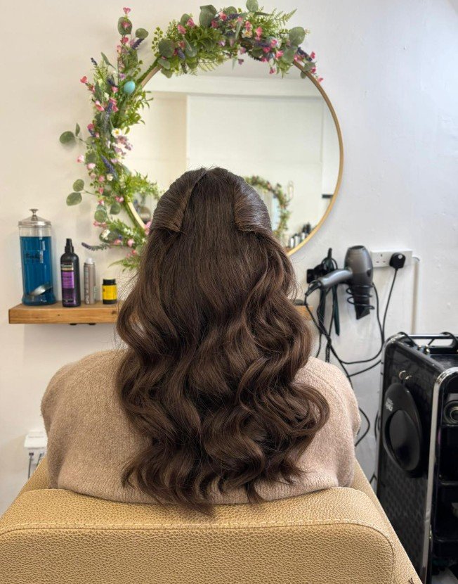 Back of a woman with long, wavy brown hair sitting in a salon chair, facing a mirror decorated with flowers and greenery.