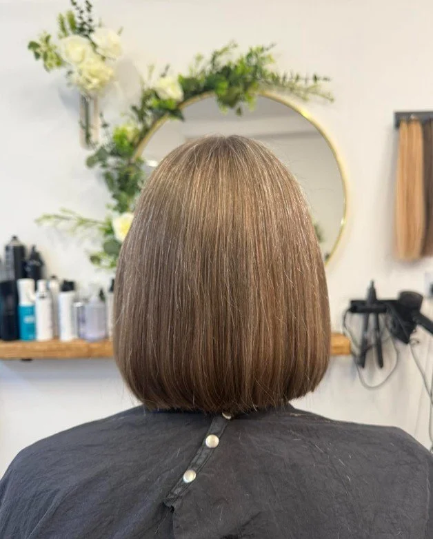 Back of a woman's head showing a freshly cut, chin-length bob hairstyle with straight, shiny, light brown hair, taken inside a salon with a round mirror, white flowers, and shelves with hair products visible.