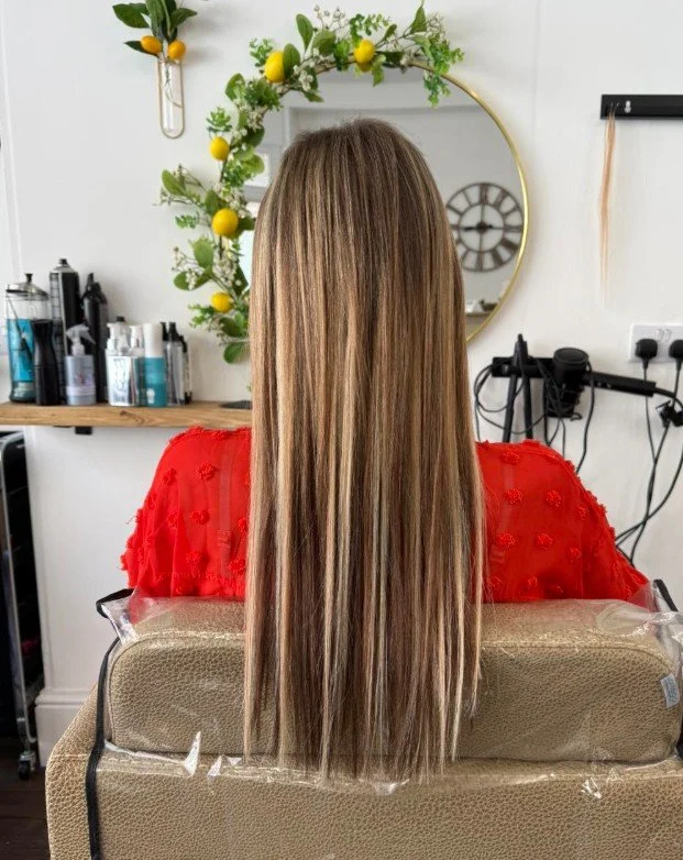 Back of a woman with long, straight, blonde hair wearing a red top, sitting in a salon chair in front of a mirror with decorative greenery and lemons, salon tools and products visible on shelves
