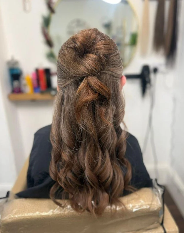 Back view of a woman with long, curled brown hair styled in a half-up, half-down hairstyle, sitting in a salon chair.