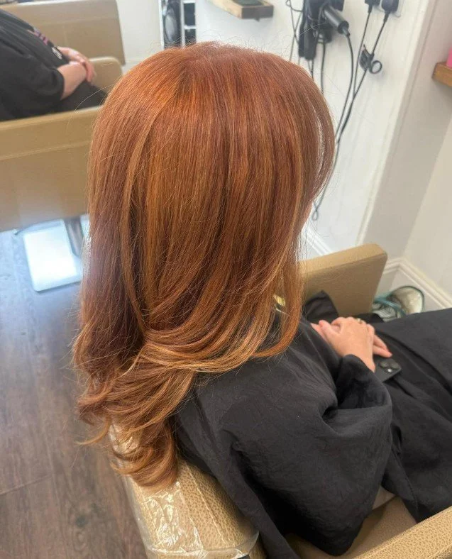A woman with long, wavy, red hair sitting in a salon chair, wearing a black cape, with her head turned away from the camera.
