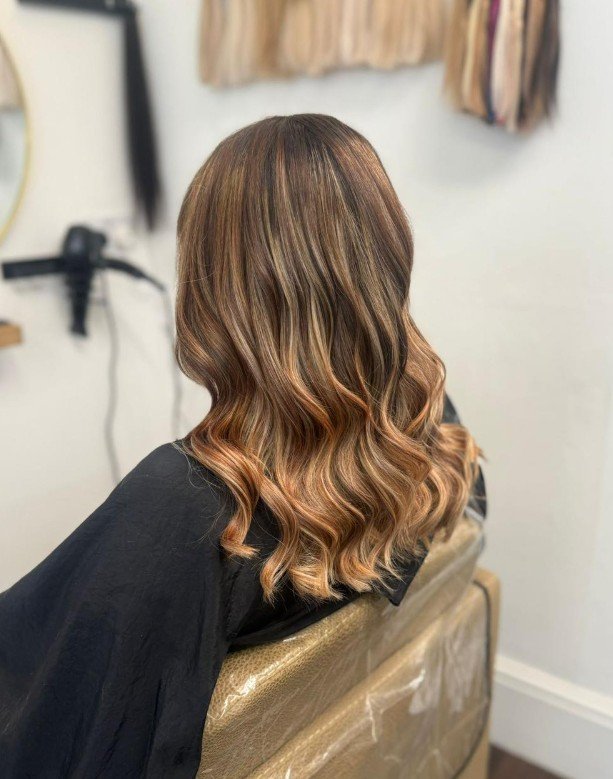 Back view of a woman with shoulder-length, wavy hair dyed in warm caramel and honey blonde highlights, sitting in a salon chair.