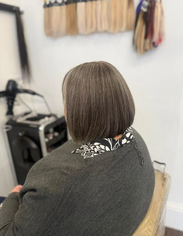 Back view of a person with a short, straight, brown bob haircut, sitting in a salon chair with a dark gray blazer and a black and white floral patterned shirt, with hairdressing tools and hanging hair accessories in the background.