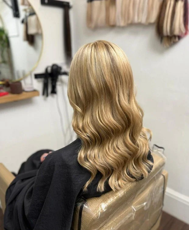 Blonde woman with long, wavy hair sitting in a salon chair, facing away from the camera.