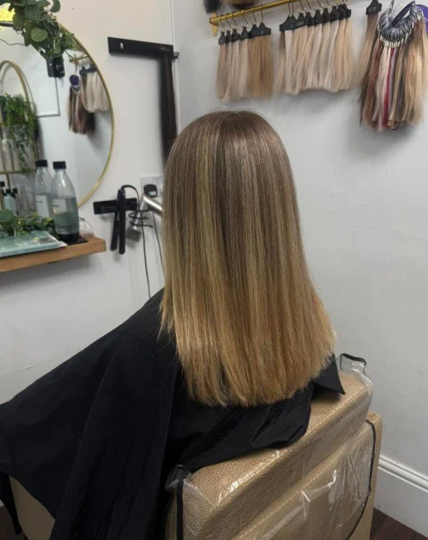 A woman sitting in a hair salon chair with freshly styled straight, shoulder-length blonde hair. The salon has a mirror, hair extensions hanging on the wall, and shelves with hair products.