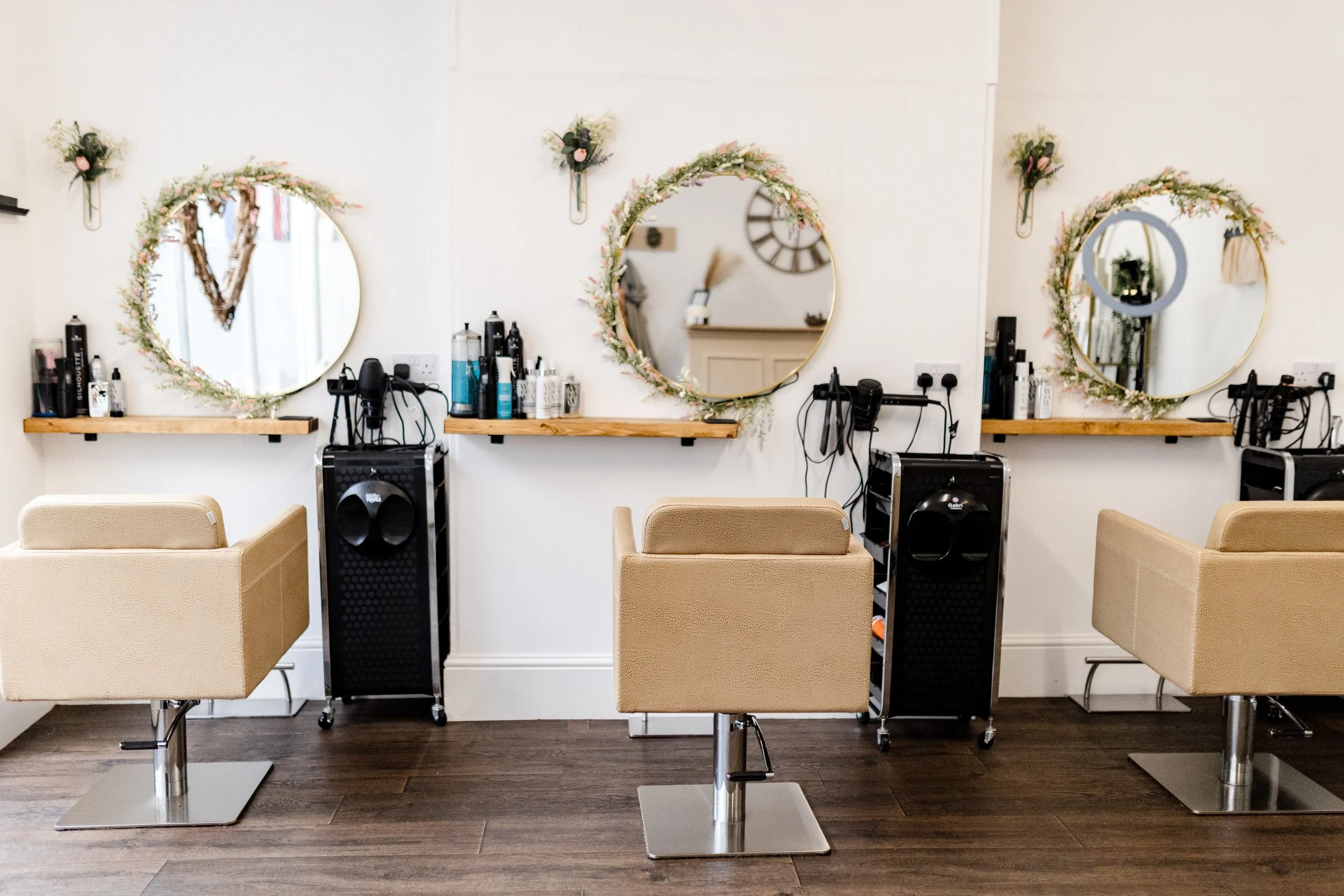 A salon station with three round mirrors decorated with floral garlands, hair styling tools, and chairs placed in front of each mirror. The station is on a dark wooden floor with beige salon chairs.