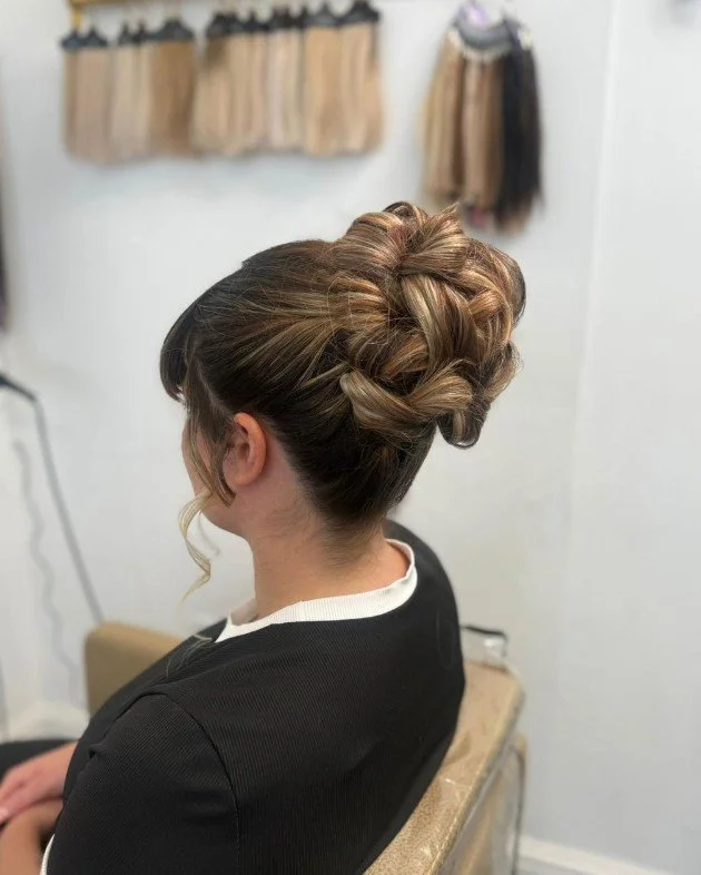 A woman with a formal updo hairstyle sitting in a salon or studio, with hair extension swatches hanging on the wall in the background.