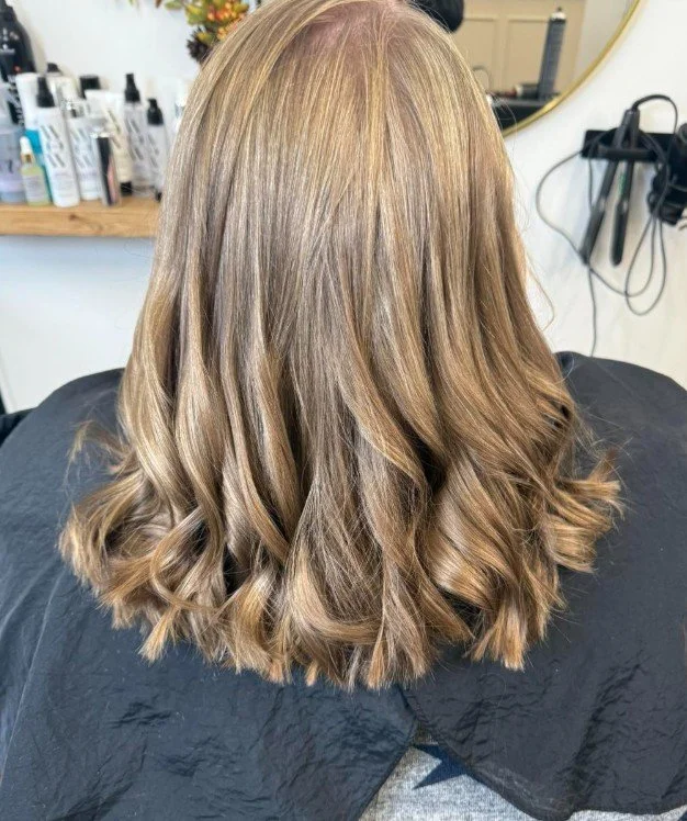 Back view of a woman with shoulder-length, wavy, blond hair in a salon setting.