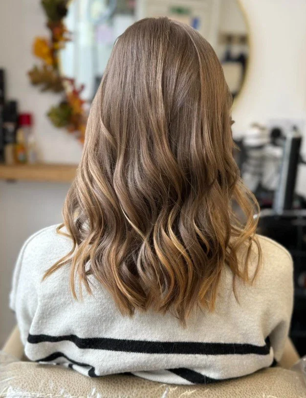 Back view of a woman with wavy, shoulder-length light brown hair wearing a light-colored sweater with black stripes in a salon or home setting.