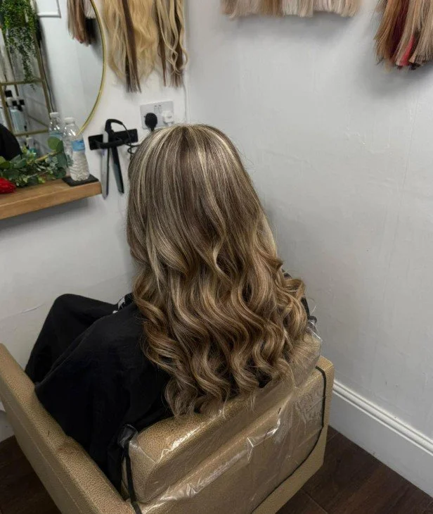 A woman with long, wavy, blonde hair sitting in a salon chair, facing away from the camera. The salon has hair samples hanging on the wall and a mirror with a small table holding water bottles and decorative plants.