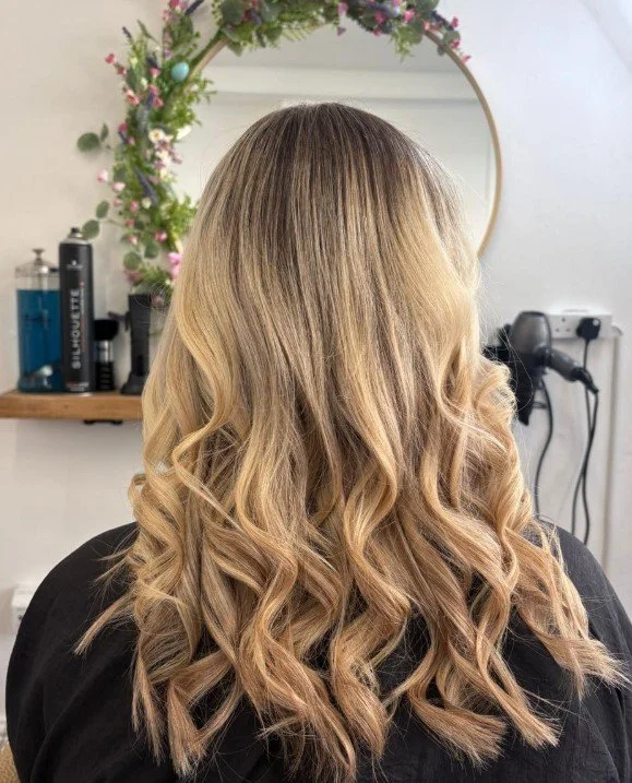 Back view of a woman with long, blonde, wavy hair, sitting in a hair salon with a mirror decorated with pink and purple flowers in the background.