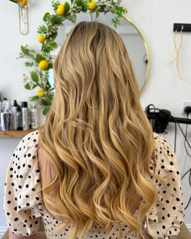 Back view of a woman with long, wavy blonde hair in a salon, featuring a decorative mirror with lemon-themed greenery in the background.