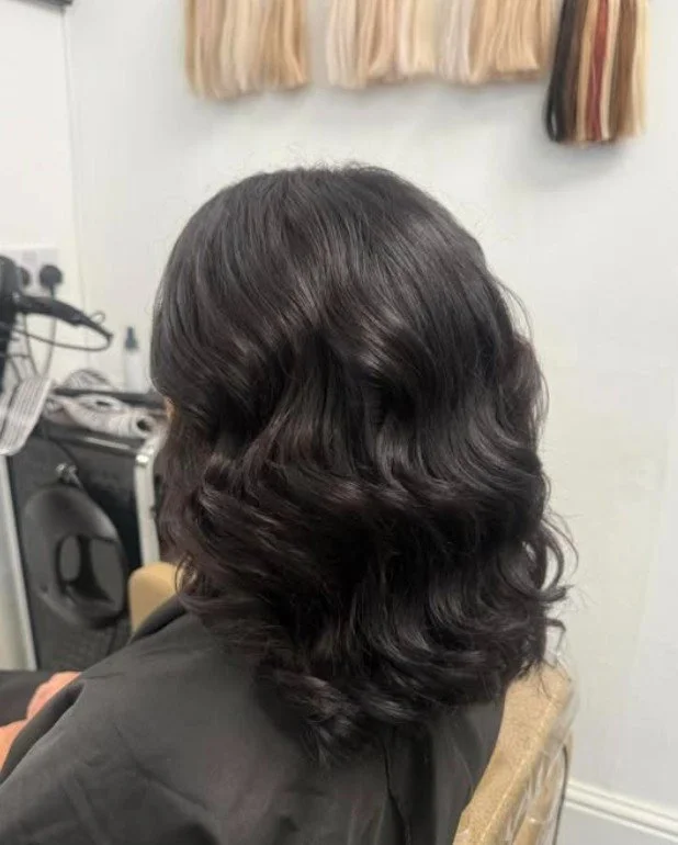 Side view of a woman with shiny, wavy dark hair sitting in a salon chair.