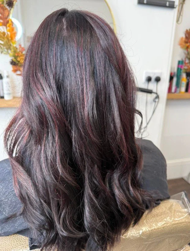 Close-up of a woman with long, wavy, dark hair with reddish highlights, sitting in a salon.