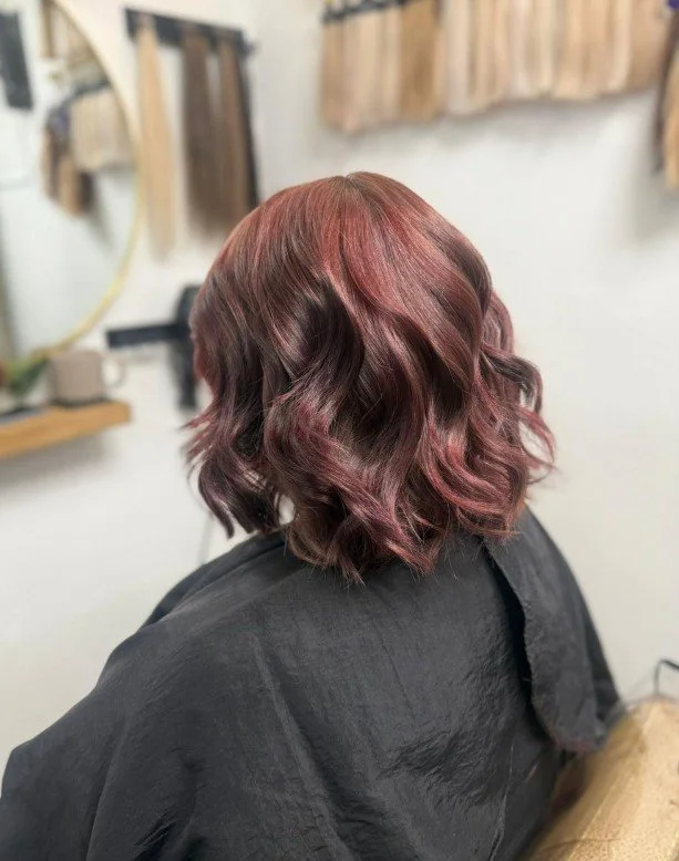 Woman with shoulder-length, wavy hair dyed in shades of red and purple, sitting in a hair salon wearing a black cape.