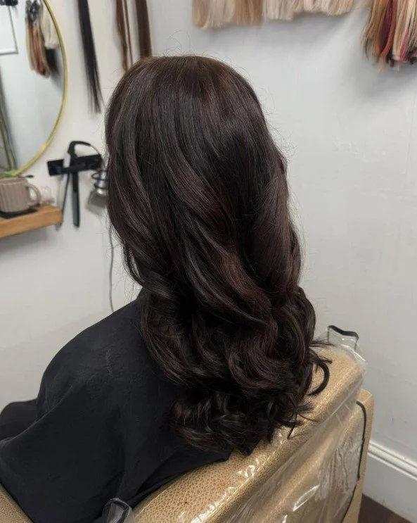 A woman with long, wavy dark brown hair seated in a salon chair with a black cape. The salon has hair tools and decorations on the walls.